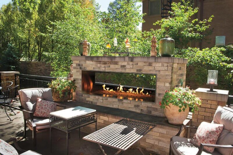 brick outdoor gas fireplace with patio furniture and greenery